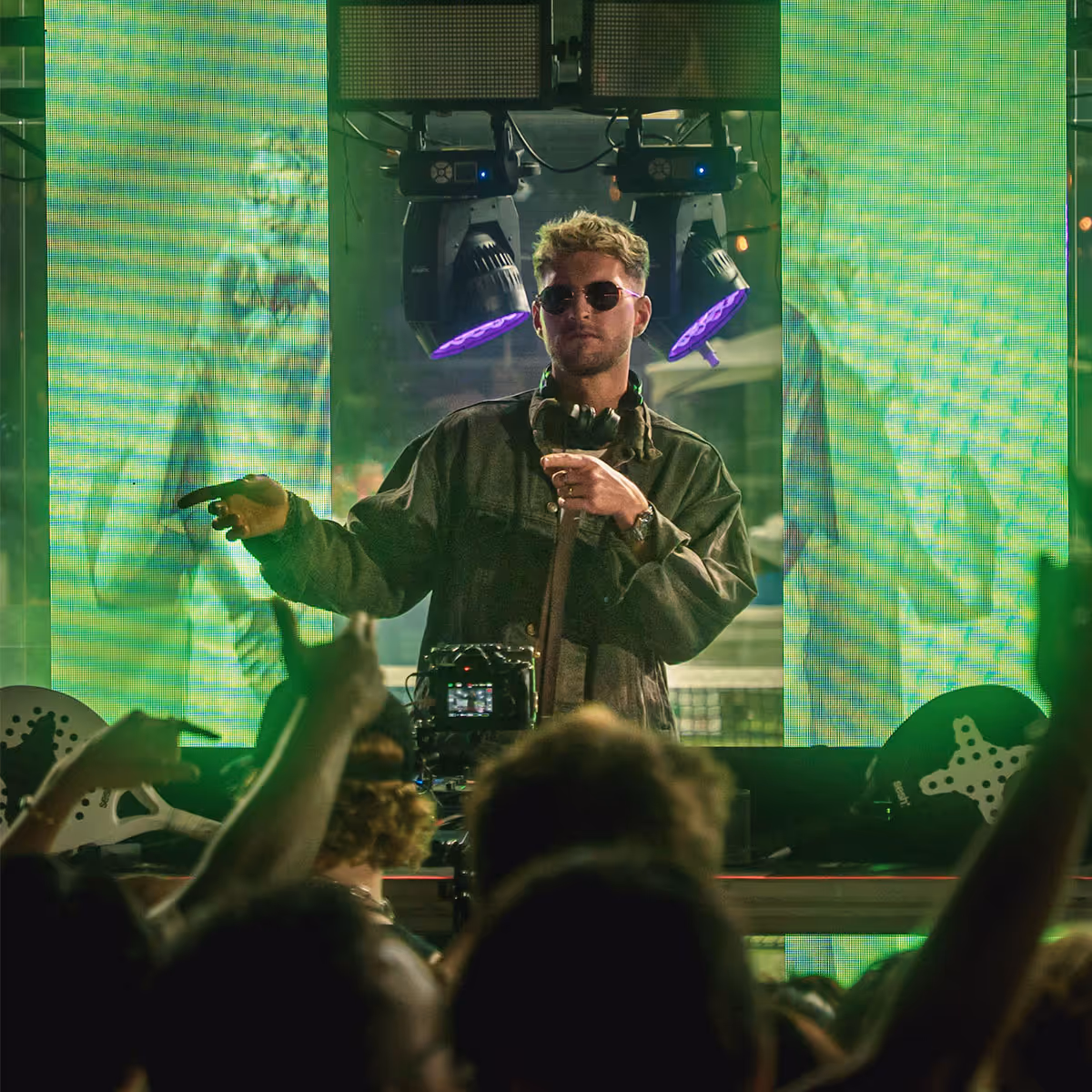 Man wearing sunglasses and headphones stands behind DJ equipment in front of a green LED screen with crowd in foreground.