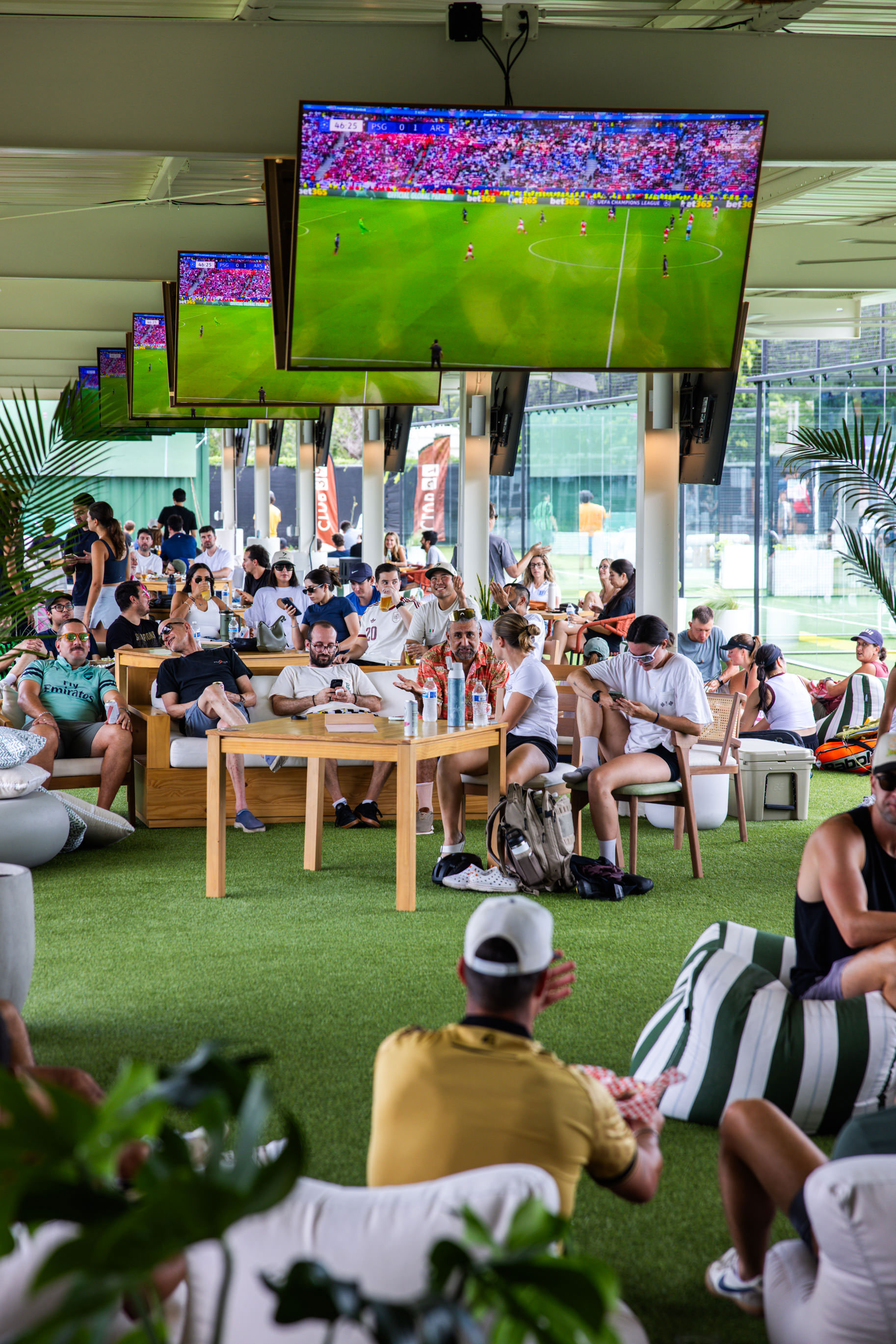 People watching a soccer match on multiple ceiling-mounted TVs in a lively indoor lounge with green carpet.