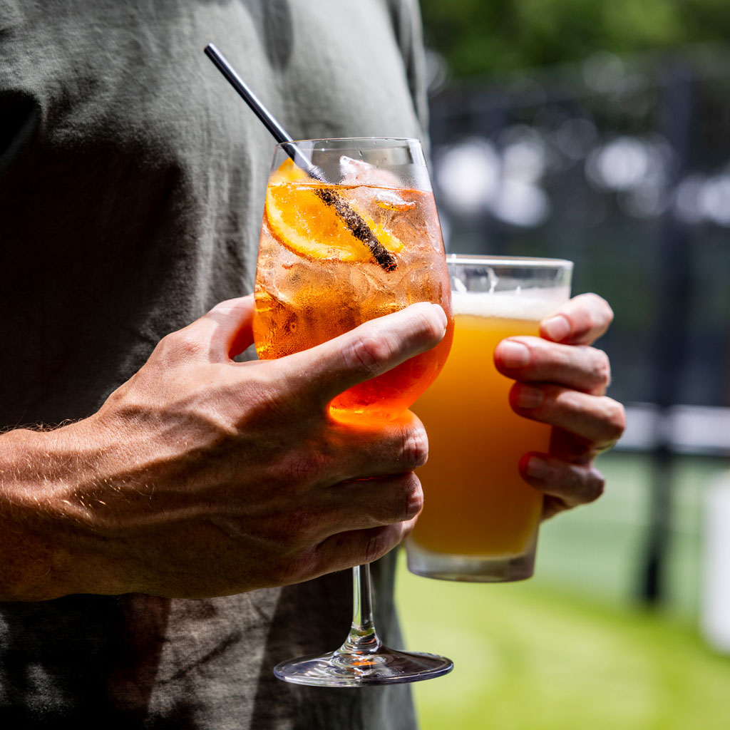 Two hands holding an orange iced cocktail with a slice and a glass of light beer outdoors.