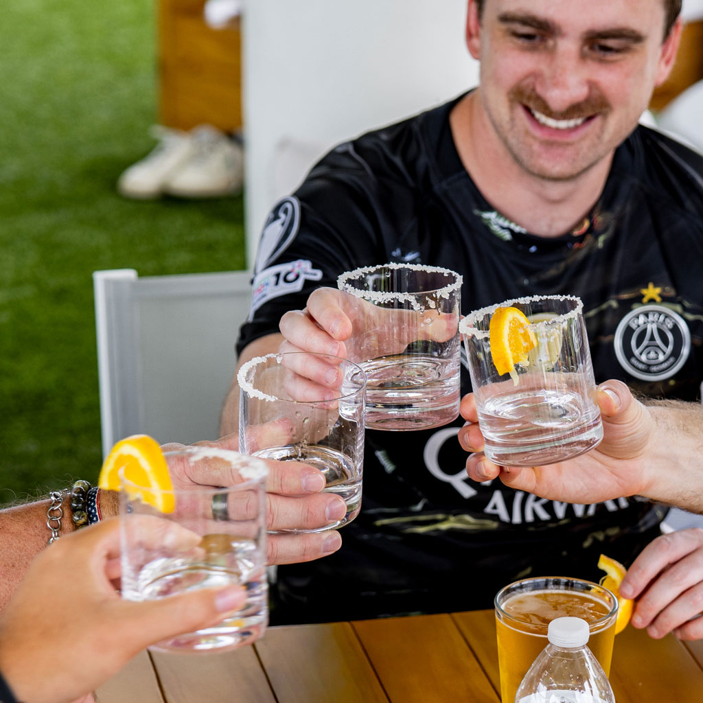 Smiling man and others clinking salt-rimmed glasses with orange slices sitting at a table outdoors.