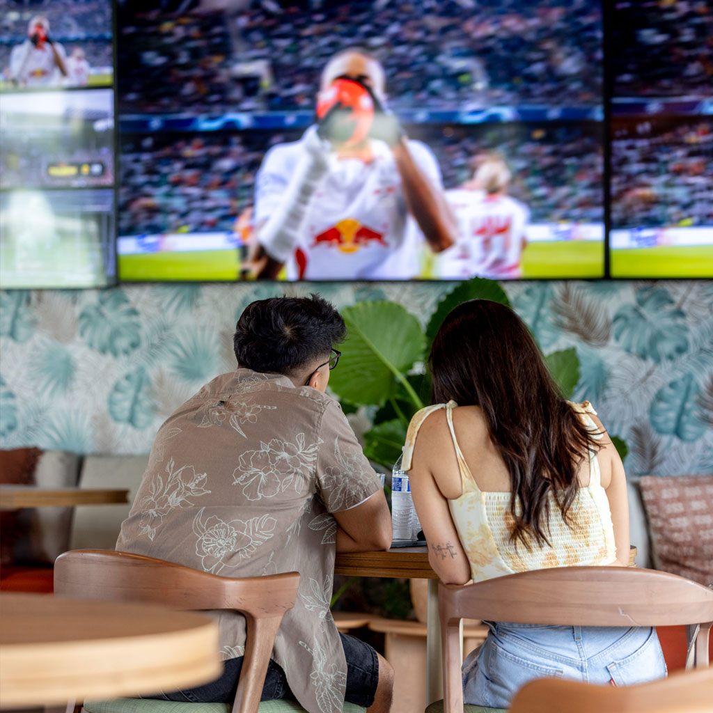 Couple seated inside bar watching a soccer game on large TV screens with green plant decor.