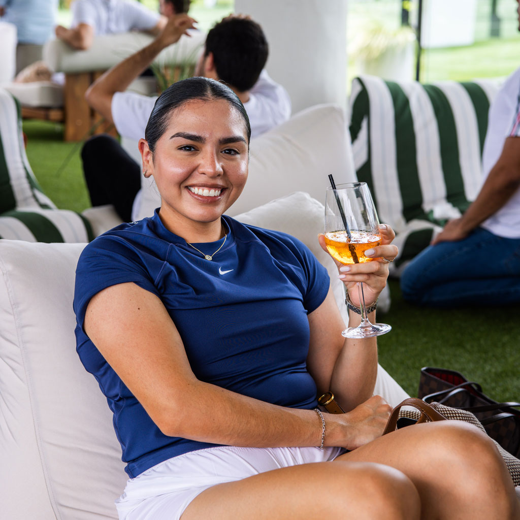 Smiling woman in navy shirt sitting on white couch holding a glass with an orange drink and straw.