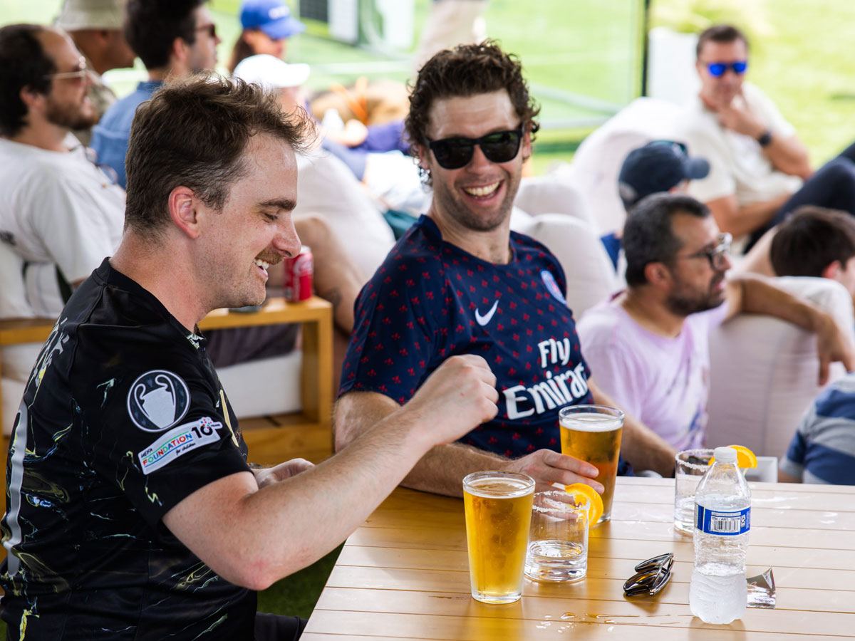 Two men smiling and sitting at a table with glasses of beer and a water bottle outdoors.