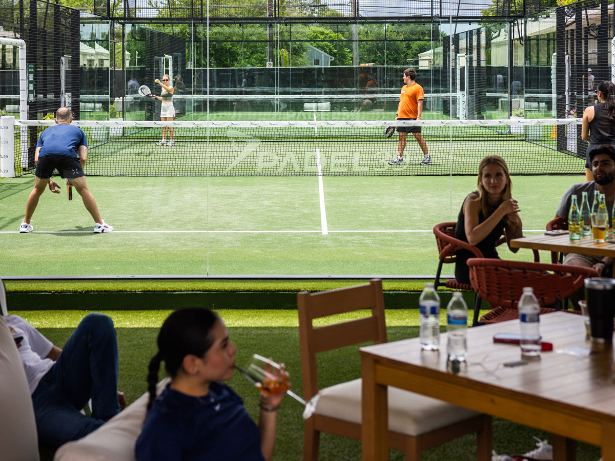 People playing padel on a court while others relax and drink nearby at outdoor seating.