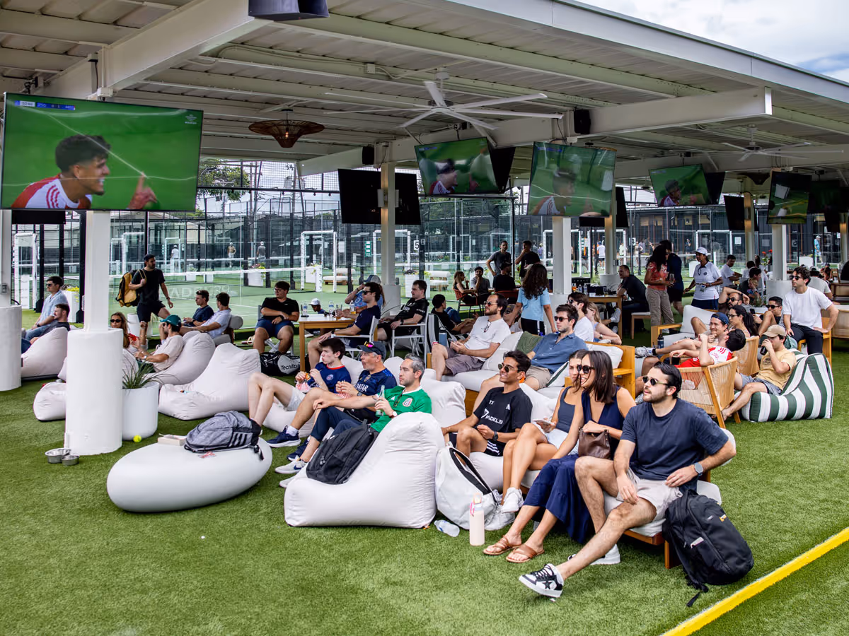 People sitting on bean bags and chairs watching soccer on multiple TVs and LED wall in a covered outdoor area.