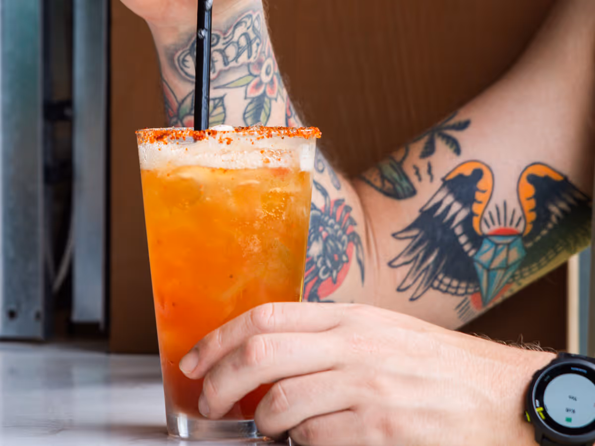 Person with tattooed arm holding a glass of iced orange drink with a chili rim and black straw.
