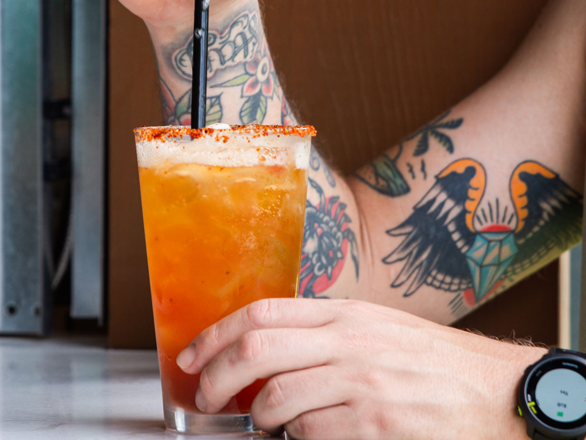 Person with tattooed arms holding a glass of iced orange drink with a chili-rimmed glass and black straw.