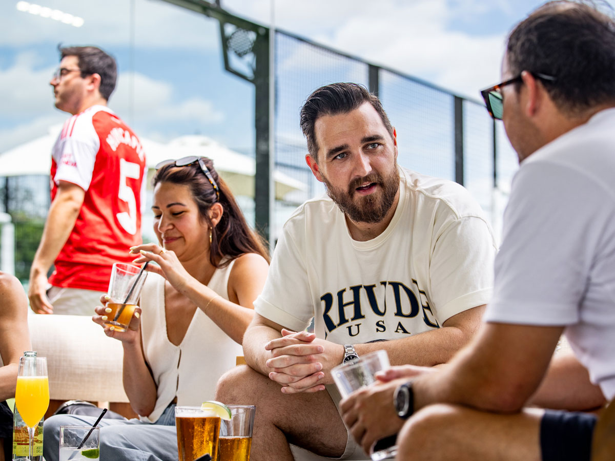 Group of friends outdoors drinking and chatting on a sunny day by a fenced court.