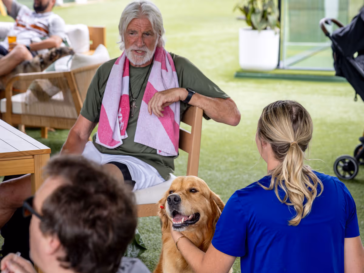 Man with white hair sitting, a woman petting a happy golden retriever at an outdoor gathering.