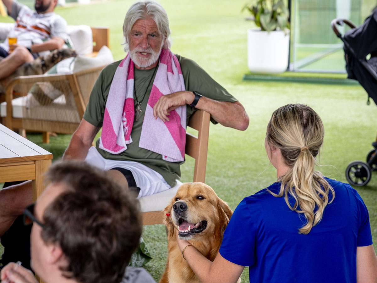 A man with white hair and a beard sitting outdoors with a dog and a woman in a blue shirt.
