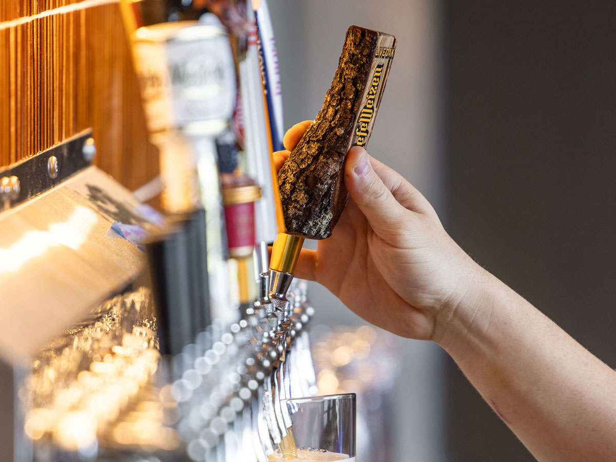 Hand pulling a beer tap handle shaped like a tree branch to pour draft beer into a glass.