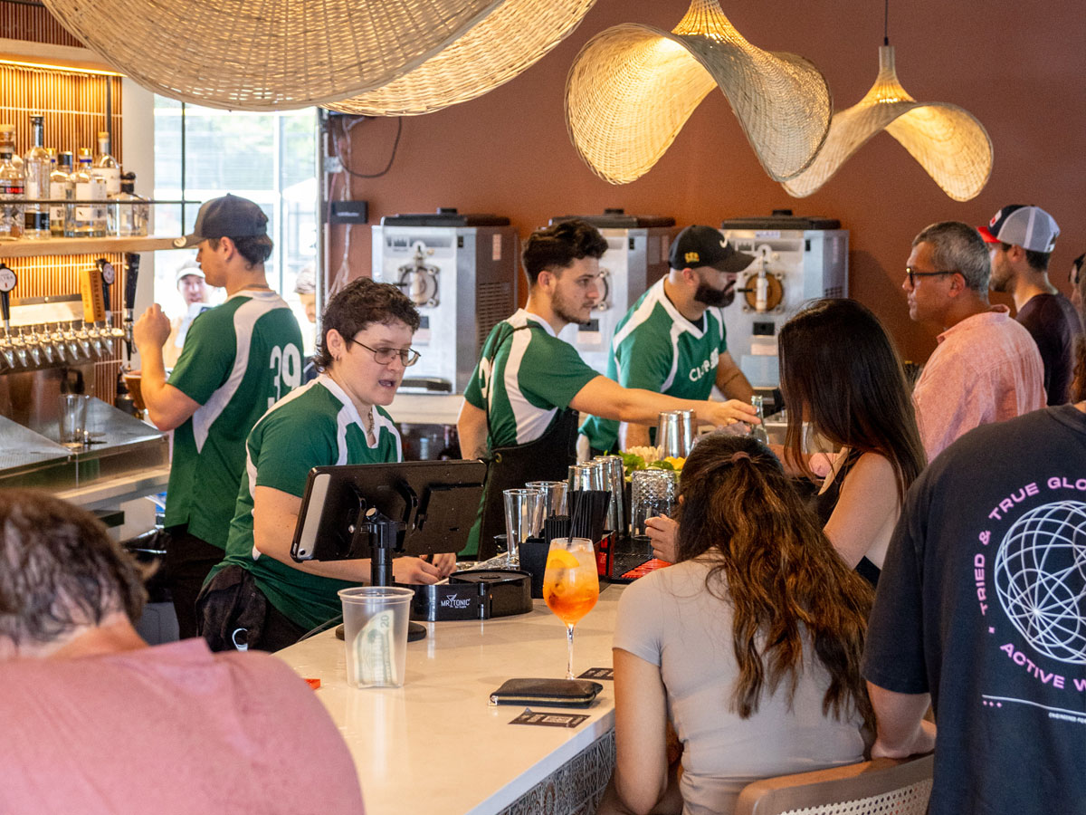 Bartenders in green shirts serve drinks to customers seated at a bar with hanging woven lights.