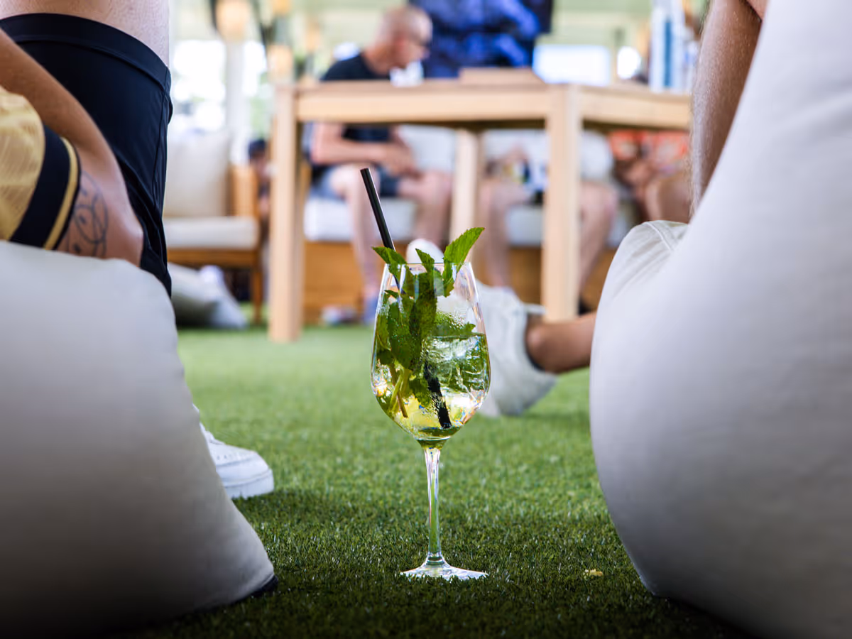 Glass of mint garnish drink with straw on turf between people relaxing on bean bags.