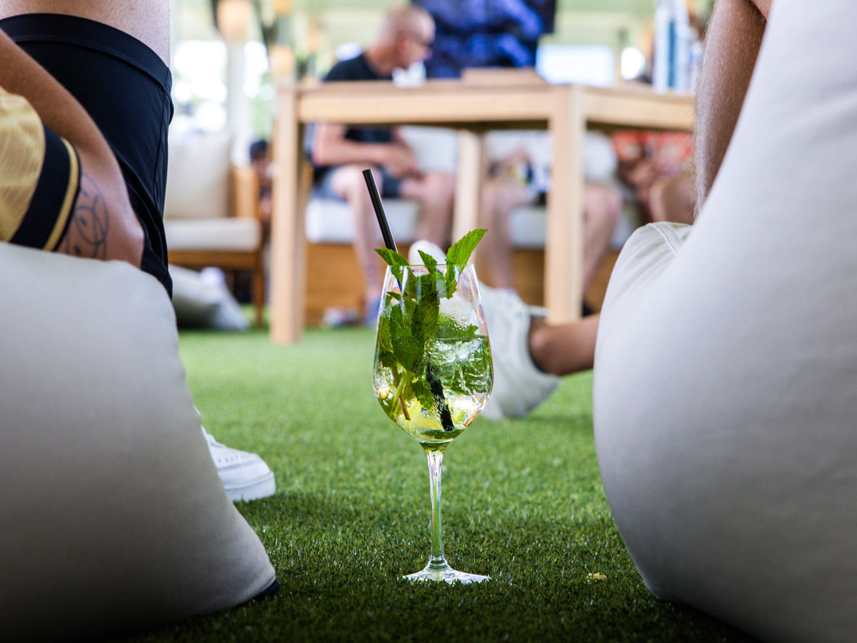 A glass of iced mint drink placed on green grass with people sitting and chatting in the background.