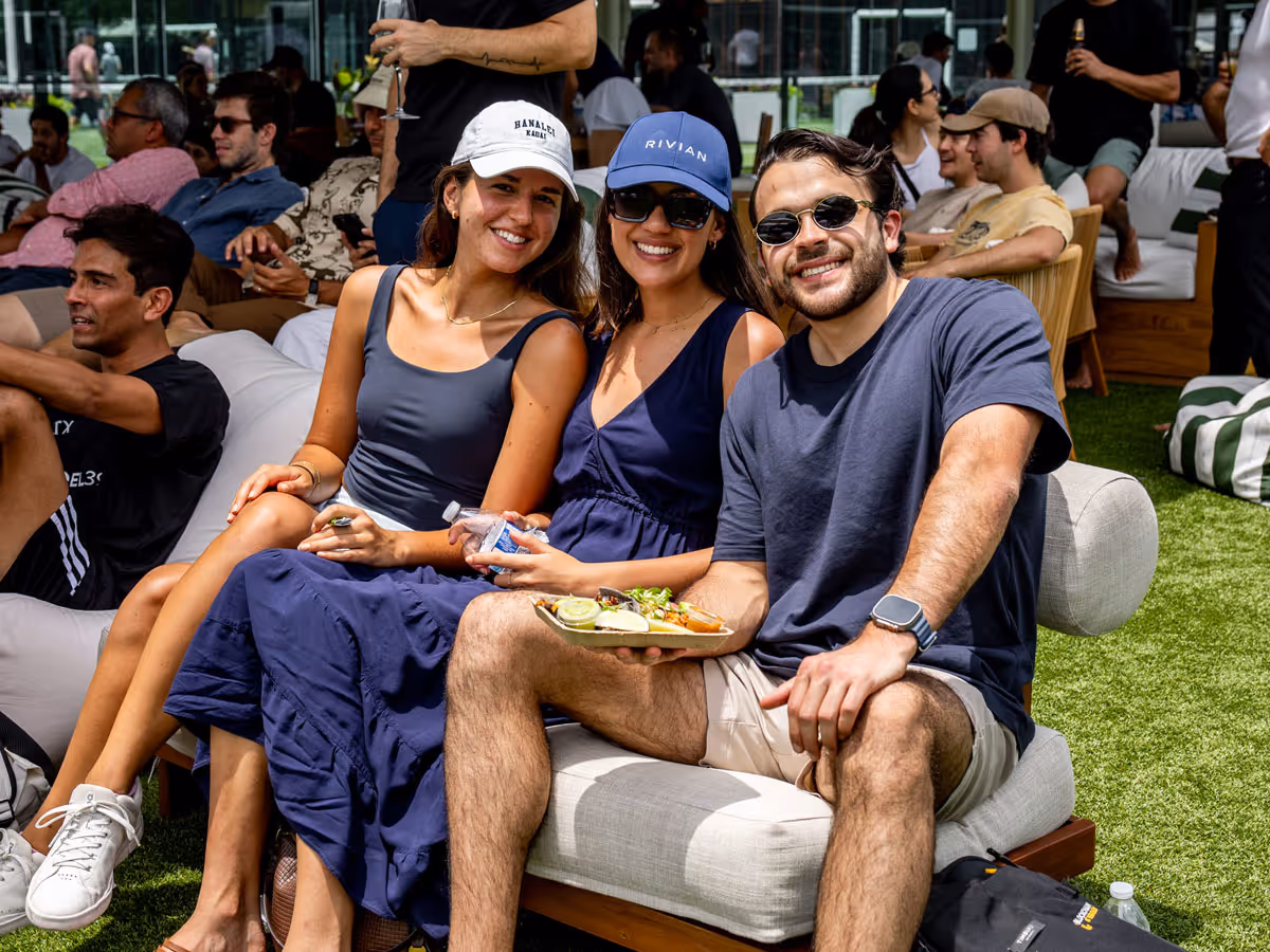 Three friends seated outdoors on a couch smiling at the camera, one holds a food plate in hand.