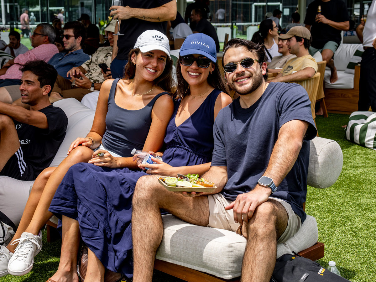 Three friends smiling outdoors on cushioned seats, one holding a plate of food and another a water bottle.