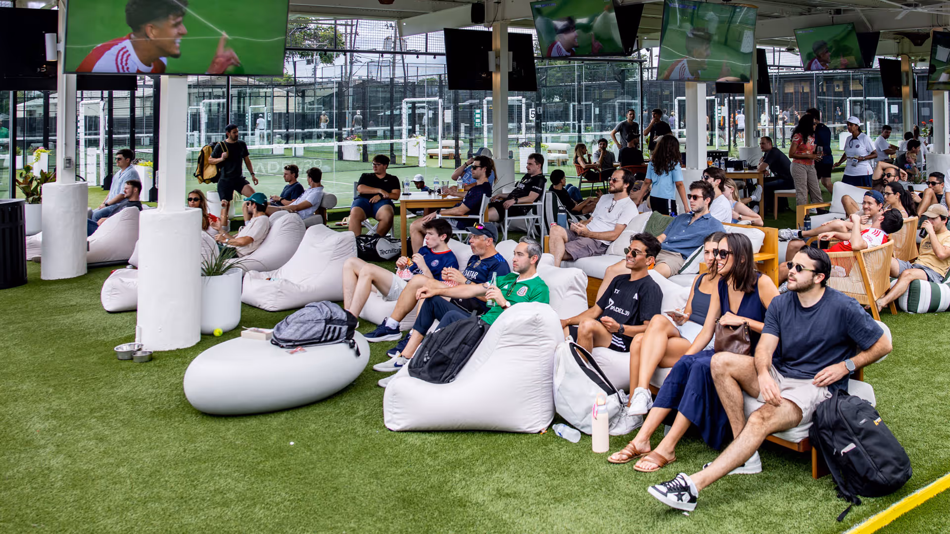 People sitting on bean bags and chairs on artificial turf watching multiple screens outdoors.