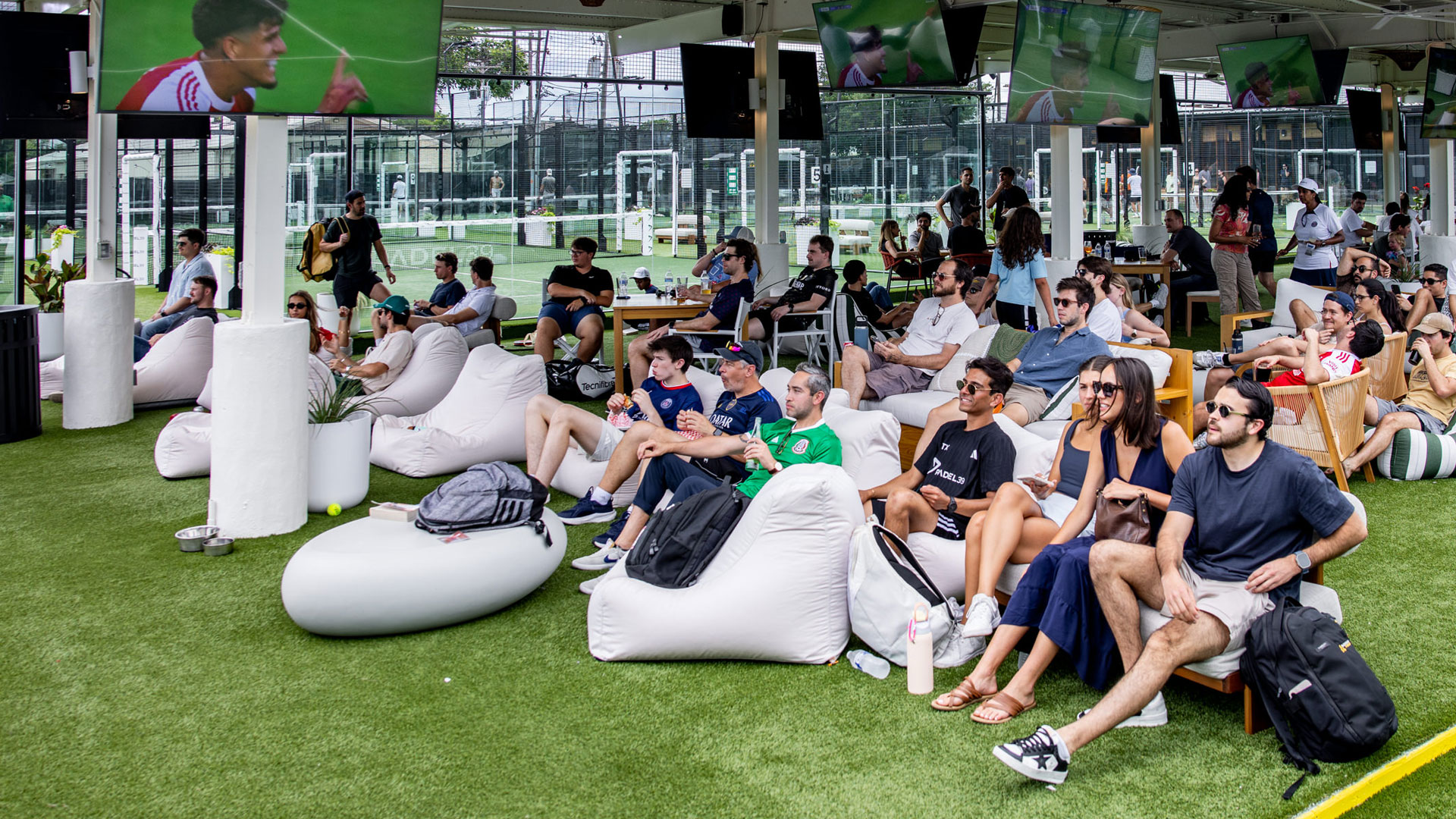 People are seated on bean bags and chairs, watching sports on a massive 20-foot LED wall and multiple TVs in an outdoor covered area.
