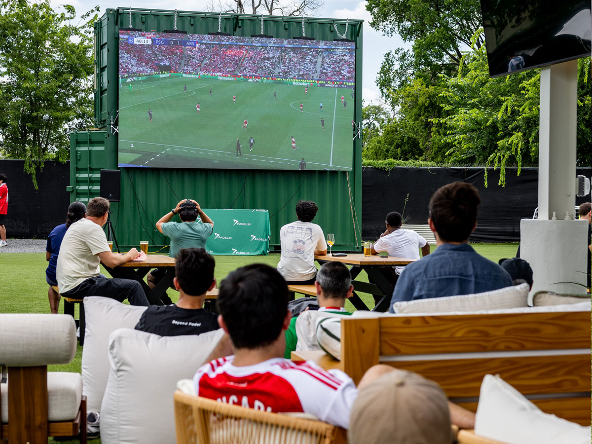 People seated outdoors watching a soccer match between PSG and Arsenal on a large screen.