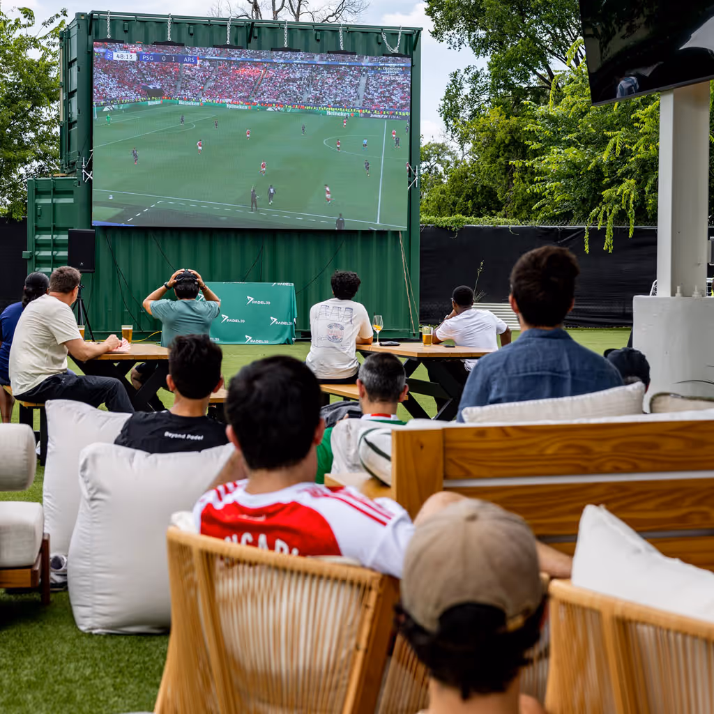People watching a soccer match on a large outdoor screen in a park-like setting.