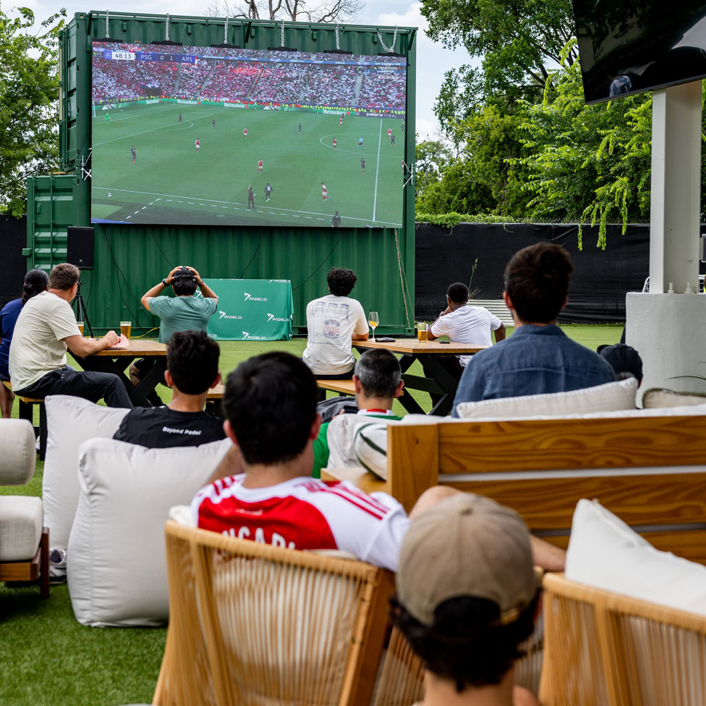 People watching a soccer match on a large outdoor screen in a park-like setting.