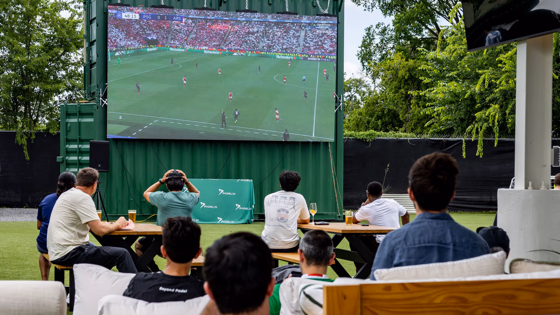 Group of people sitting outdoors watching a soccer match on a large screen with drinks on tables.