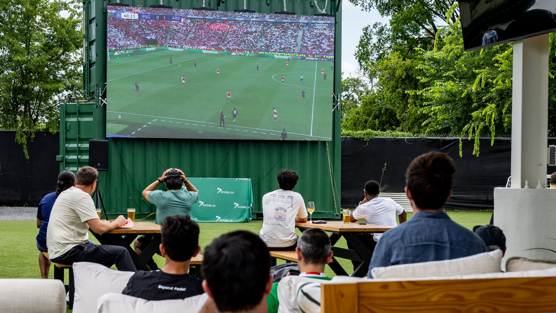 Group of people sitting outdoors watching a soccer match on a large screen with drinks on tables.