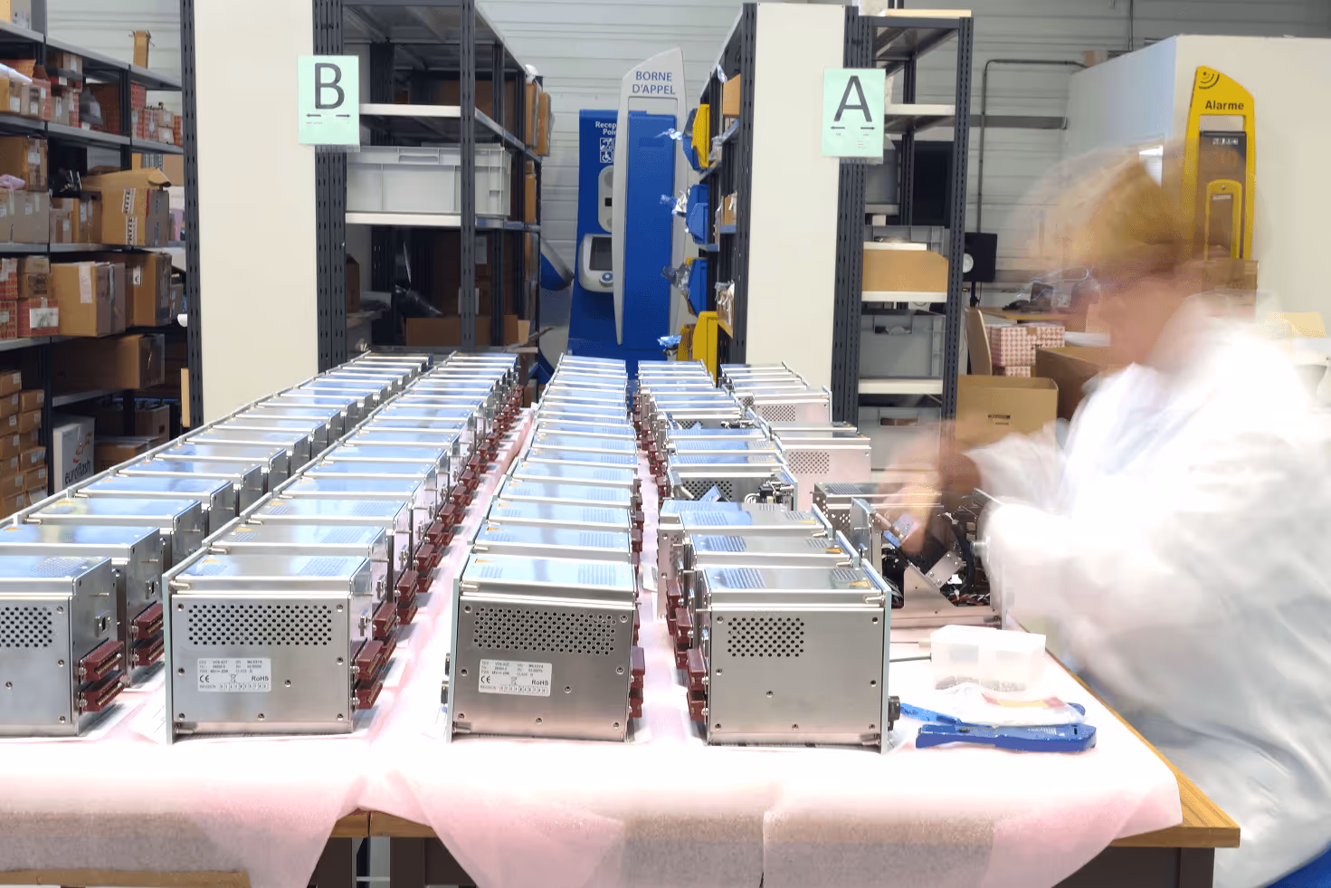 Atelier de production Milesys : rangées de modules électroniques métalliques sur banc de travail avec mousse de protection rose, technicien en blouse travaillant sur l'assemblage.