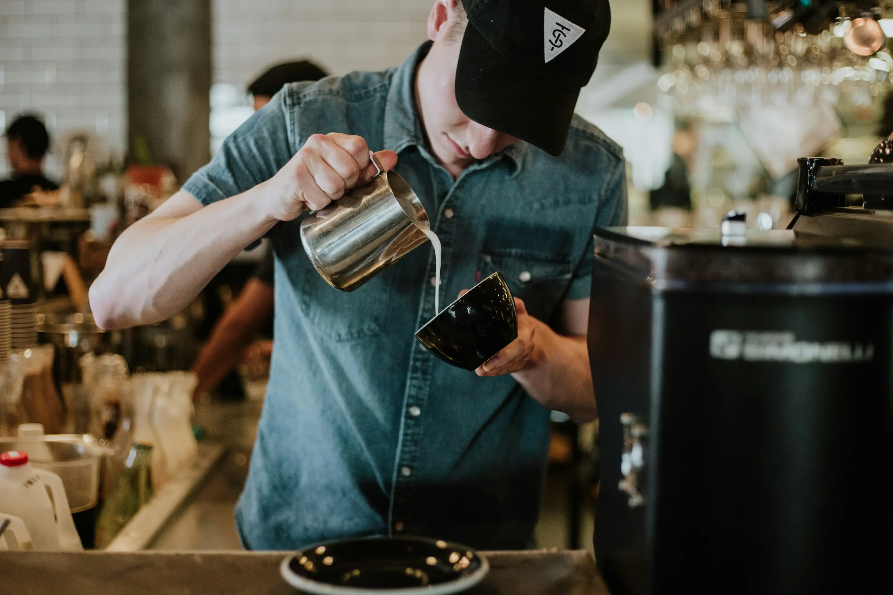 Barista in a black cap pouring milk from a metal pitcher into a black cup in a coffee shop.