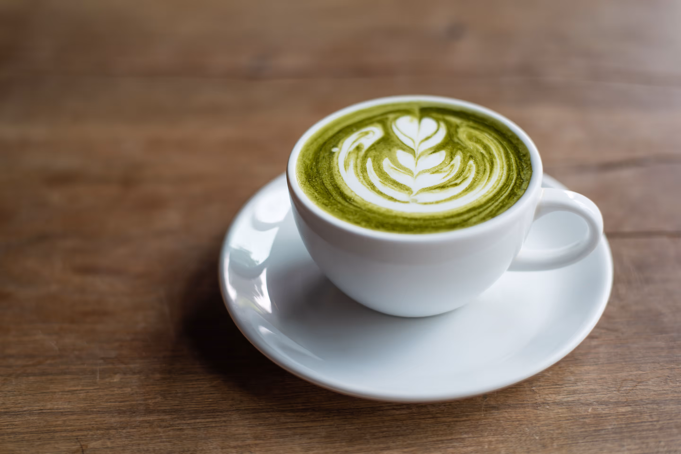 White cup of matcha latte with leaf-shaped latte art on a white saucer on wooden table.