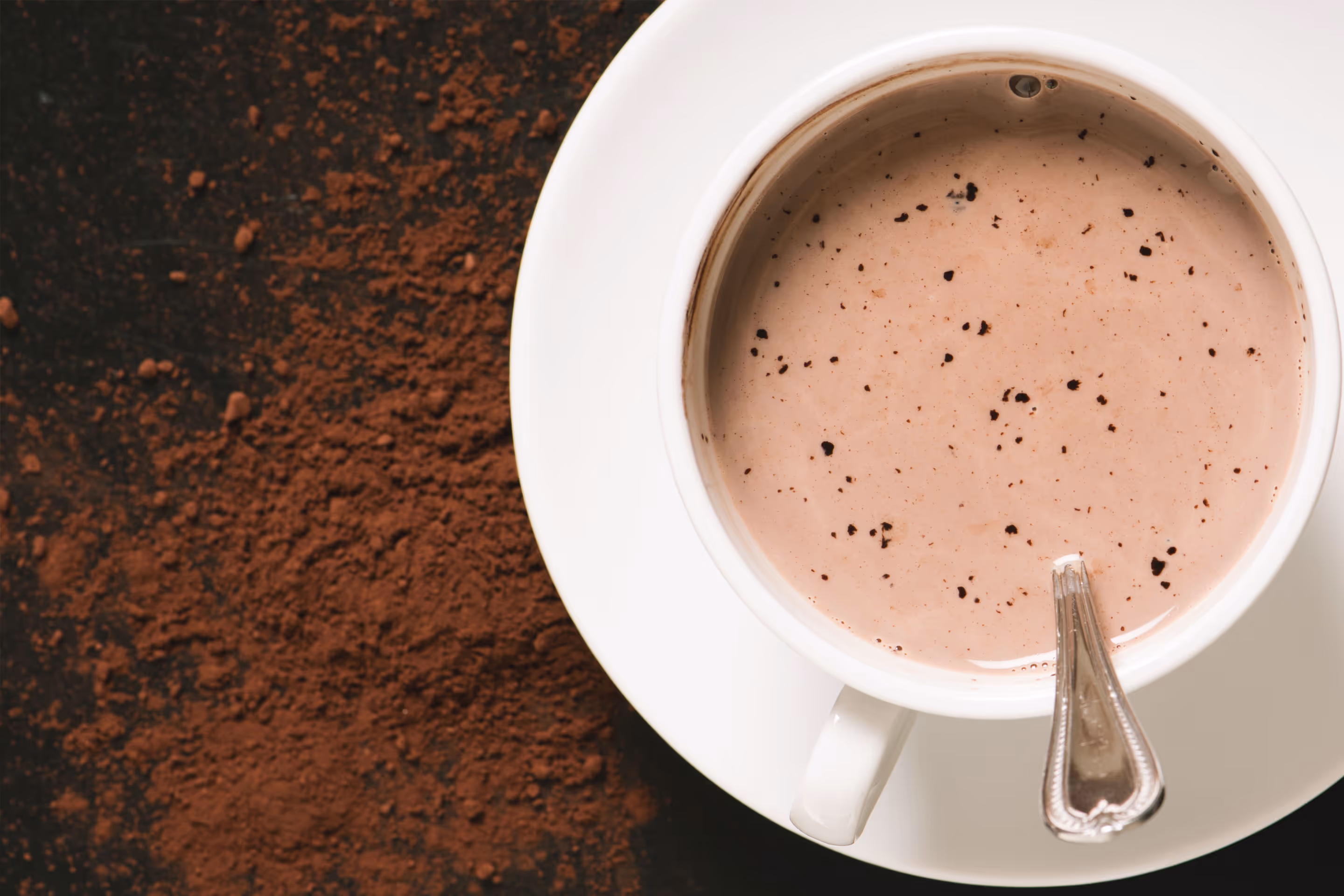 White cup of hot chocolate with chocolate flakes and spoon on saucer beside cocoa powder.