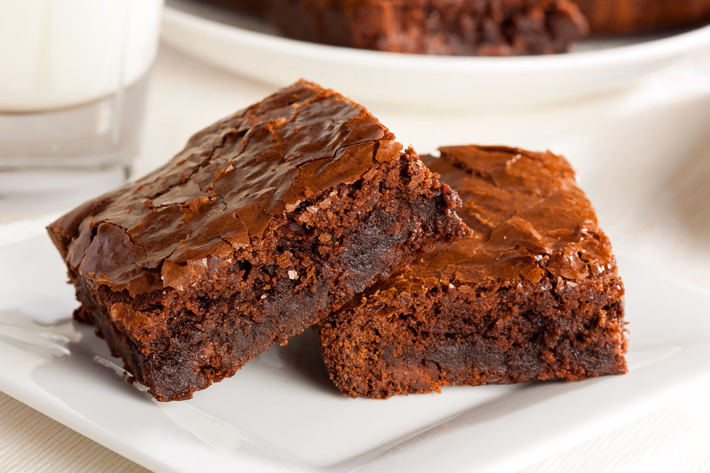 Two rich, fudgy chocolate brownies on a white plate with a glass of milk in the background.