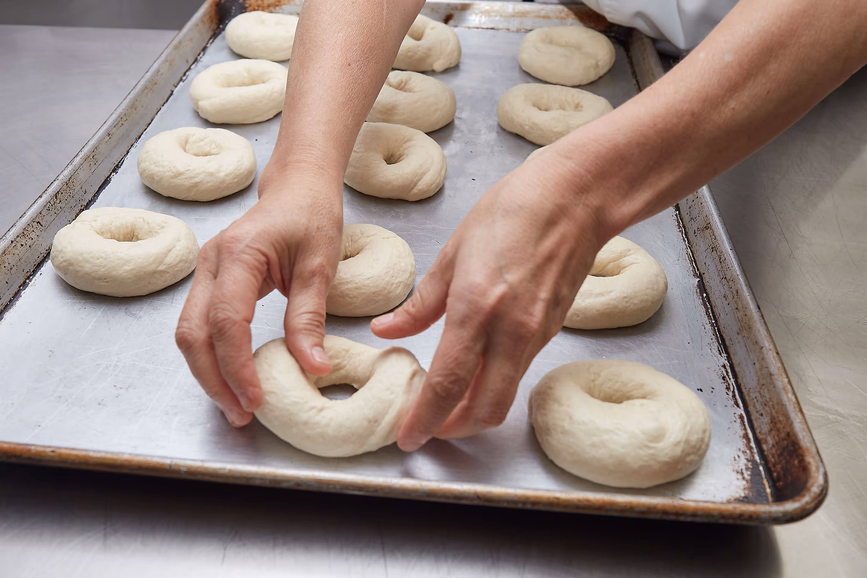 Hands shaping raw bagel dough on a metal baking sheet with multiple bagels arranged.