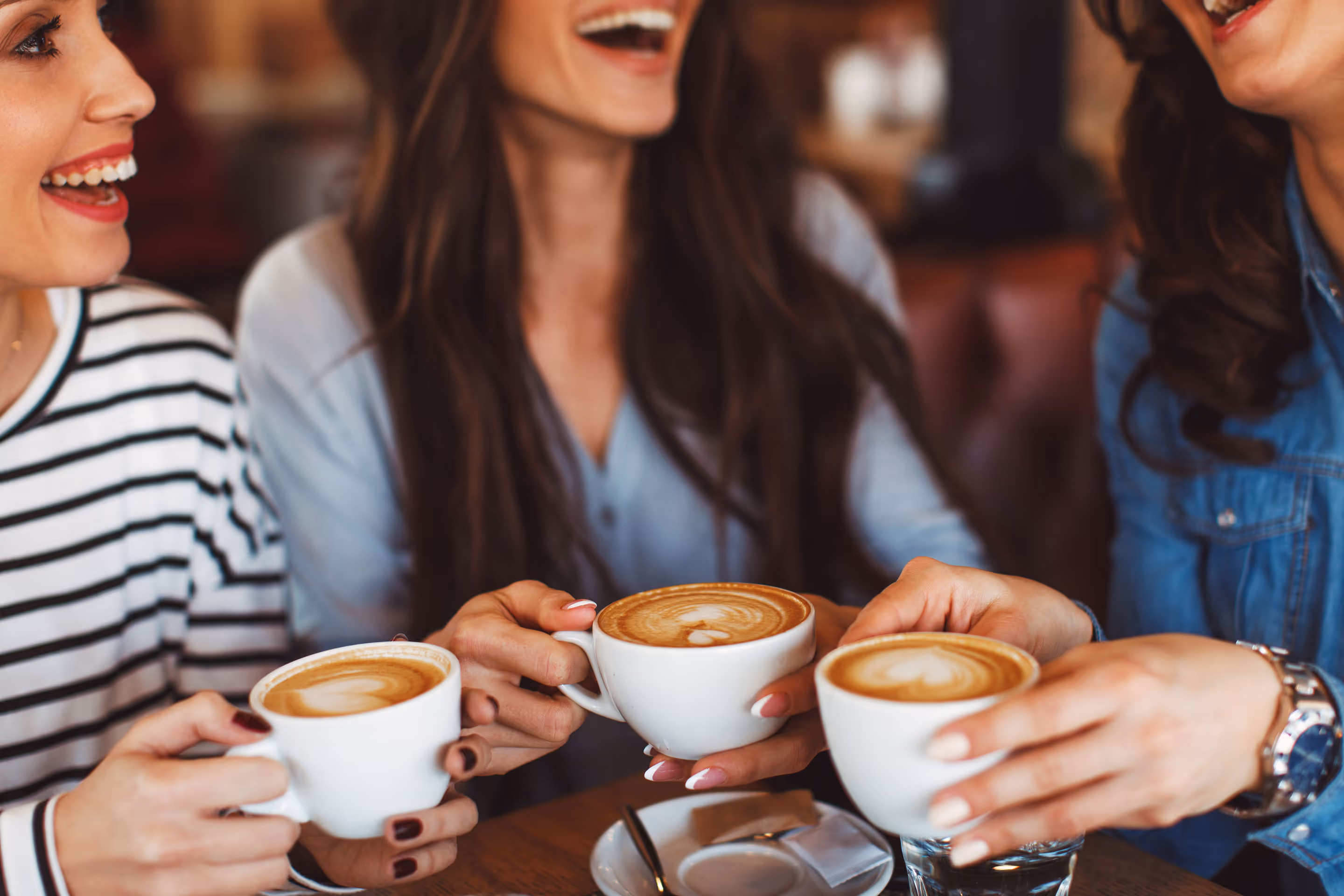 Three women laughing and holding white cups of latte with foam art in a cozy café.