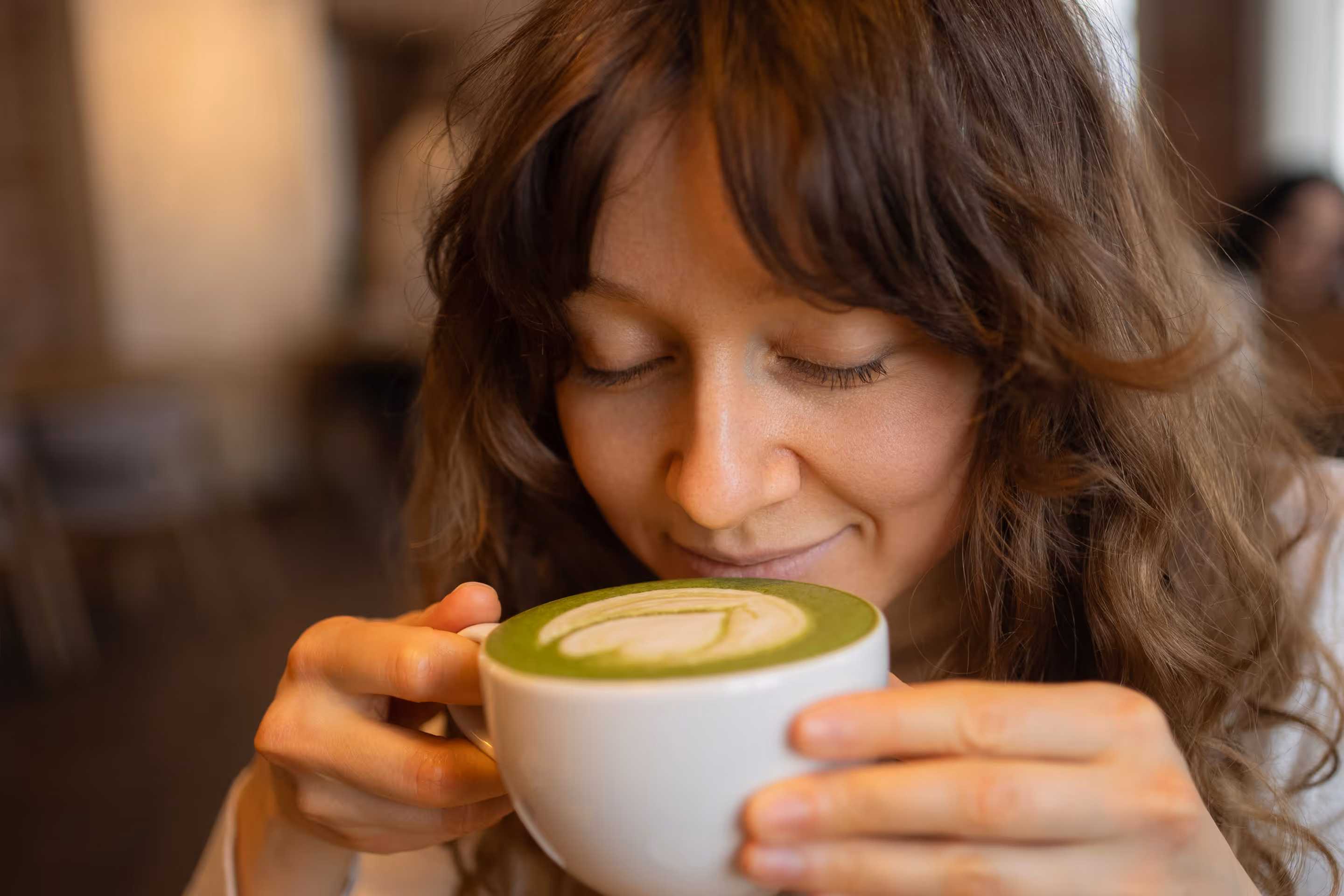 Woman with curly hair smelling a latte with green matcha foam art in a white cup.