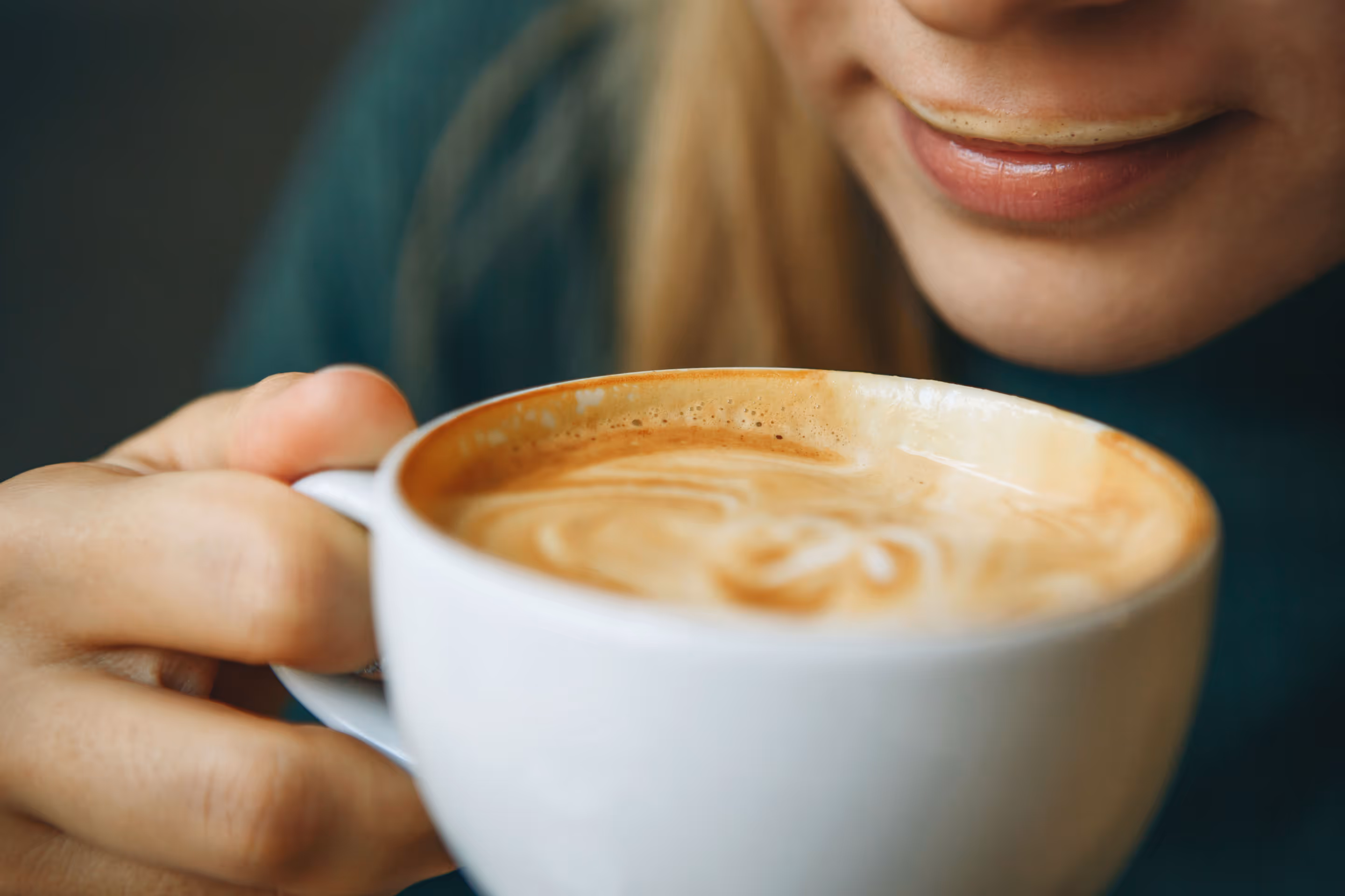 Person holding a white cup of cappuccino near their lips with a slight smile.