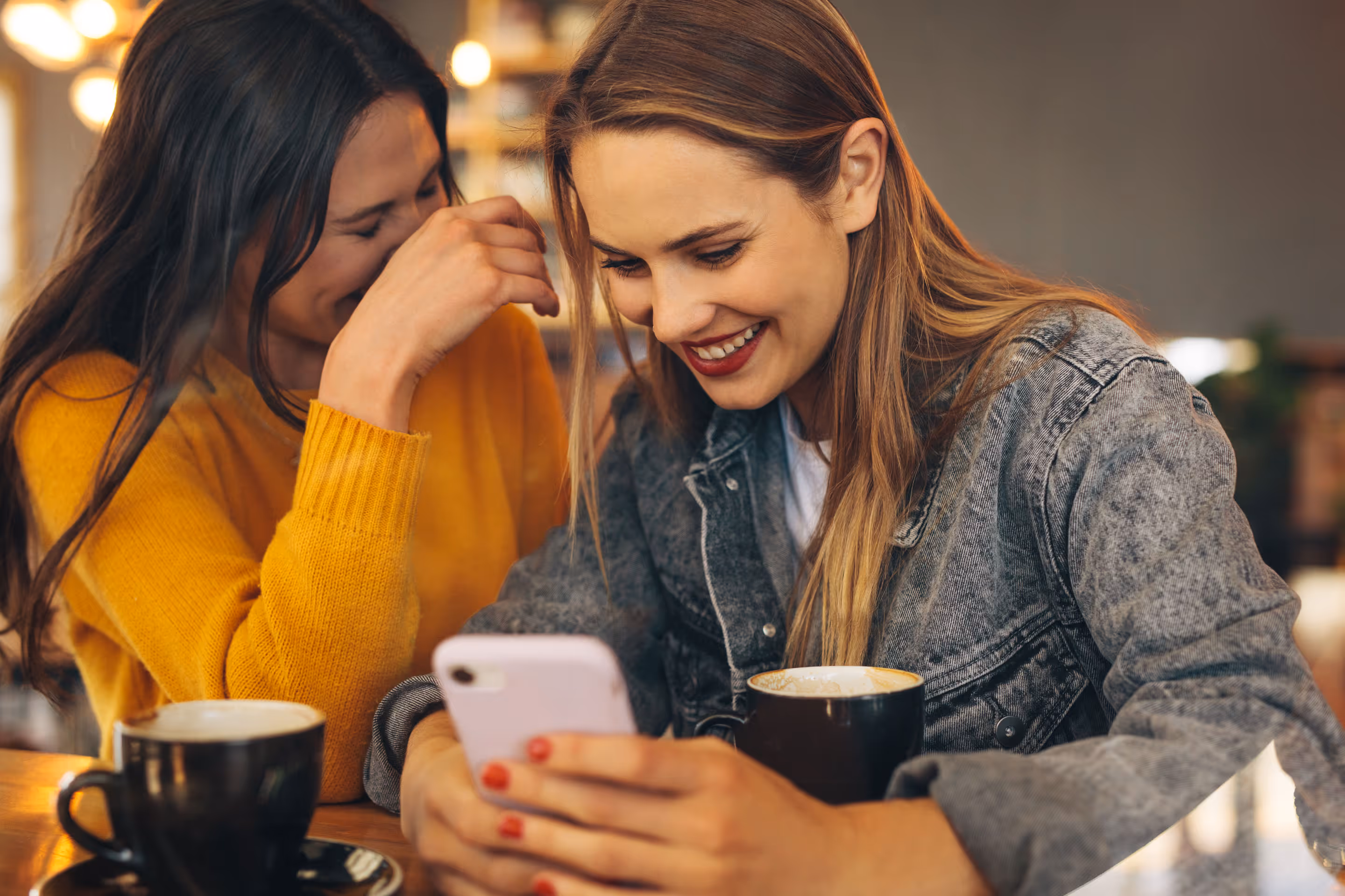 Two women laughing together in a cafe while looking at a smartphone, with coffee cups on the table.