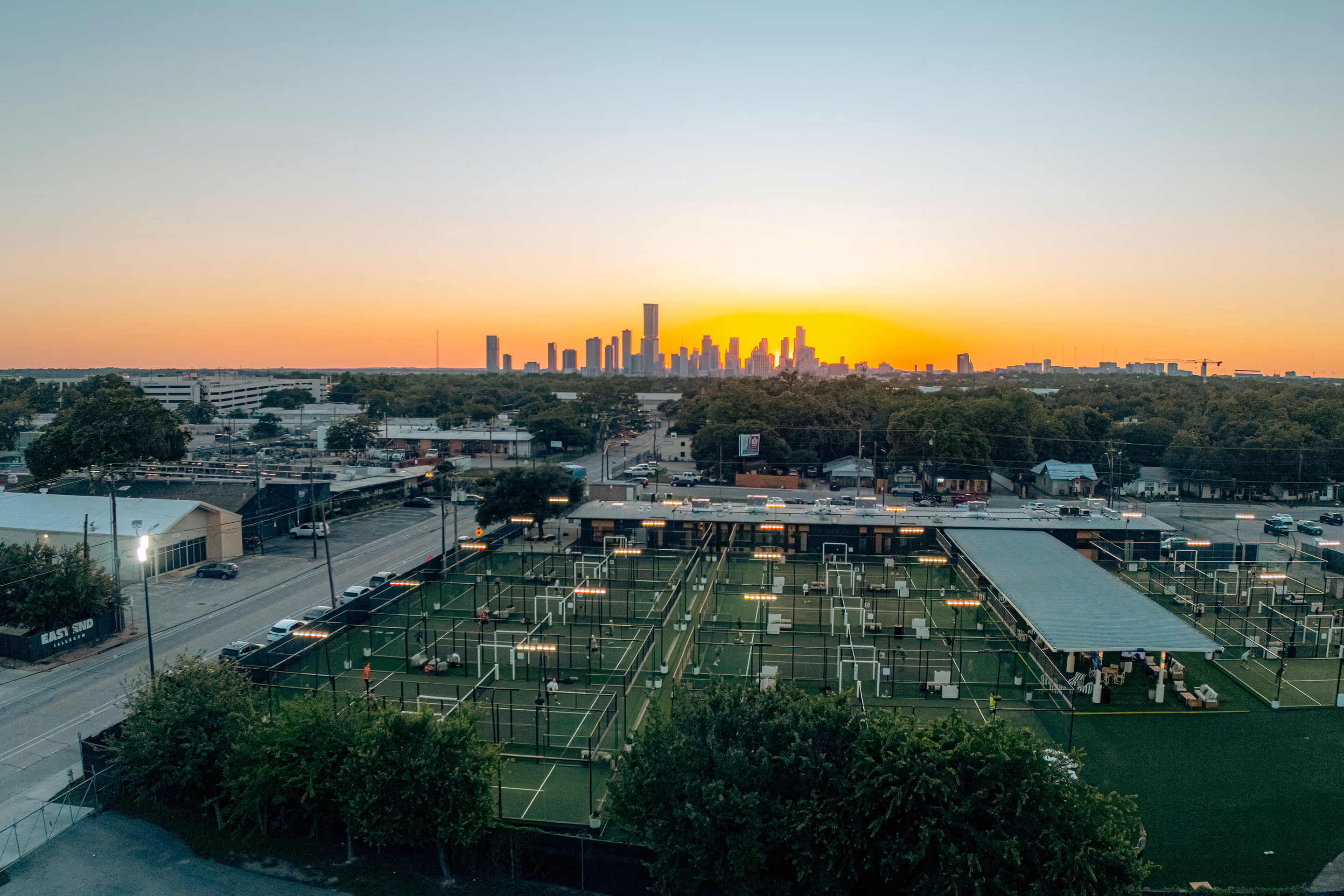 Multiple padel tennis courts with players and city skyline at sunset in the background.
