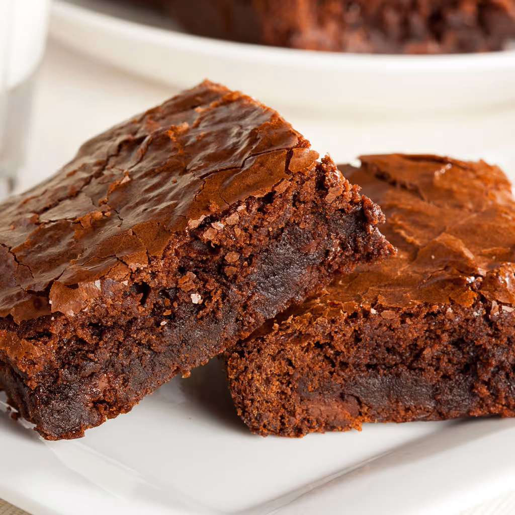 Two rich, fudgy chocolate brownies on a white plate with a glass of milk in the background.