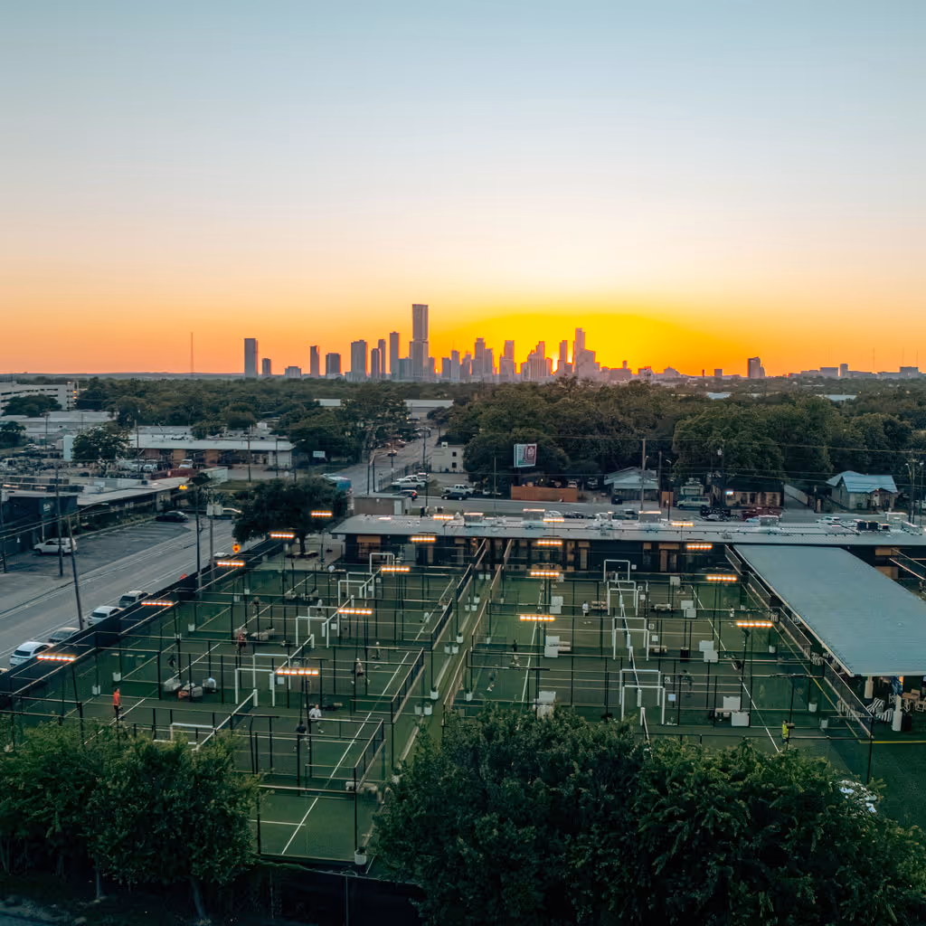 Multiple padel tennis courts with players and city skyline at sunset in the background.