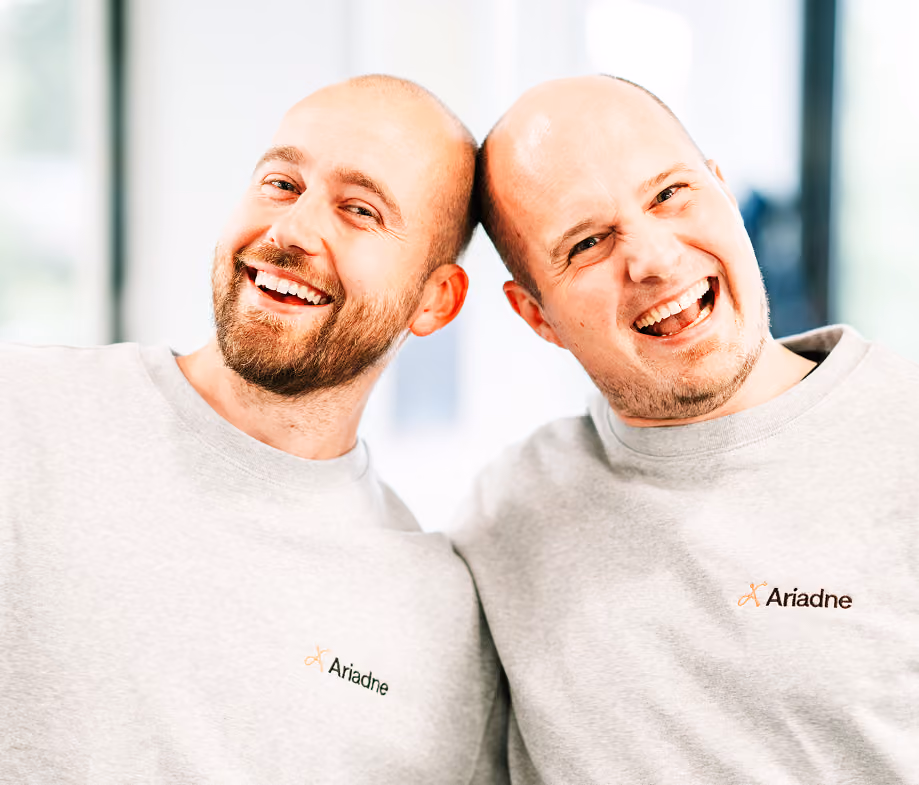 Zwei lächelnde Männer lehnen mit den Köpfen aneinander, tragen graue Ariadne-Pullover.