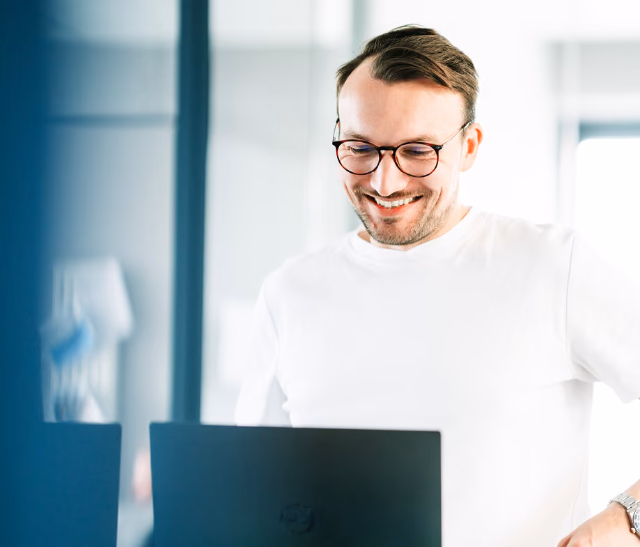 Lächelnder Mann mit Brille arbeitet an einem Laptop in einem hellen Büro.