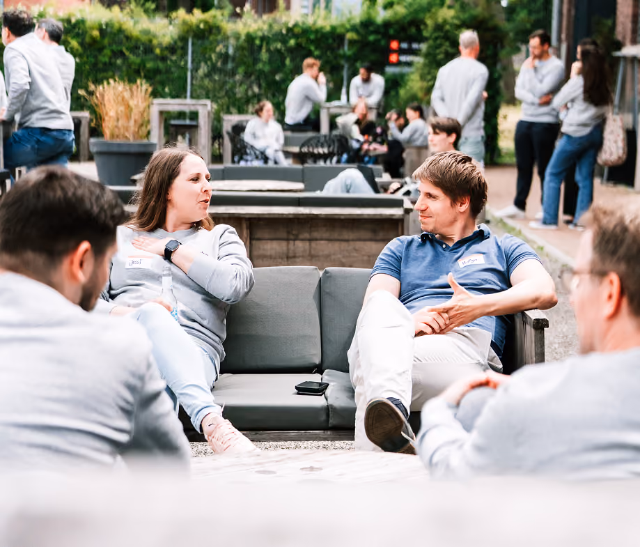 Menschen sitzen und unterhalten sich auf einer Terrasse mit Garten und Sofas im Freien.