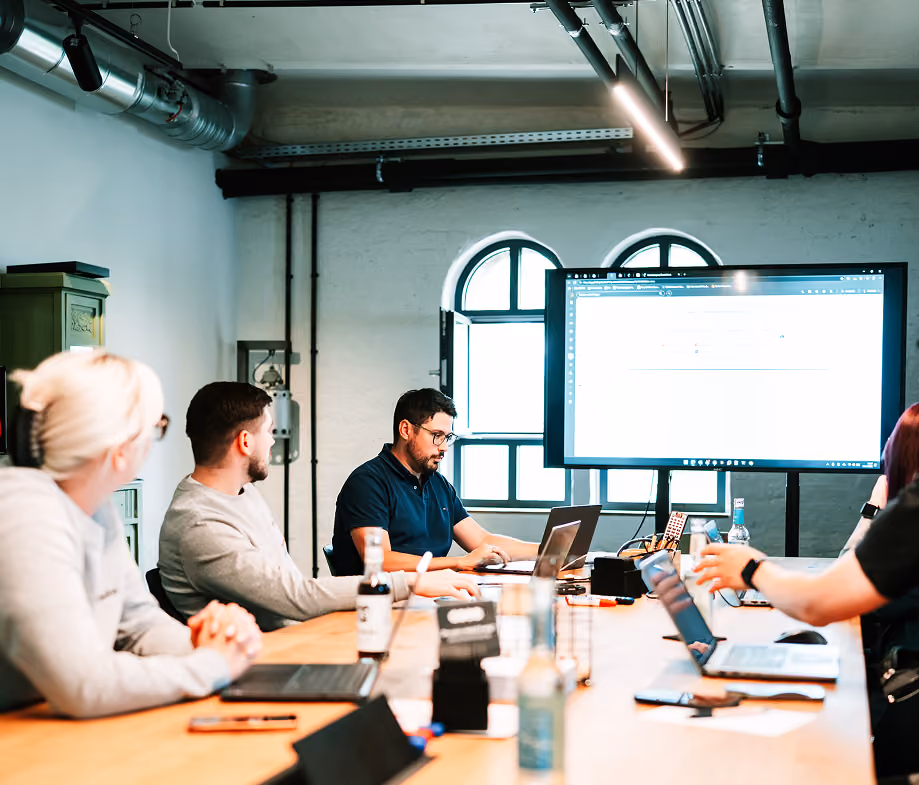 Personen arbeiten mit Laptops in einem Besprechungsraum mit großem Bildschirm und Fenster im Hintergrund.