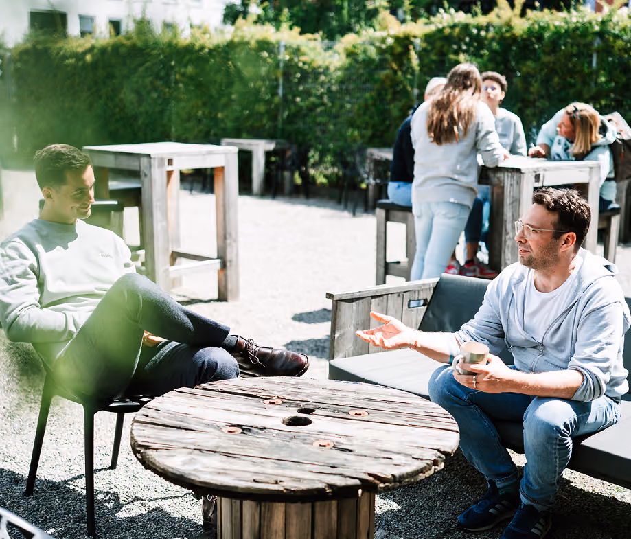 Zwei Männer unterhalten sich in einem sonnigen Außenbereich mit Holztischen und Bänken.