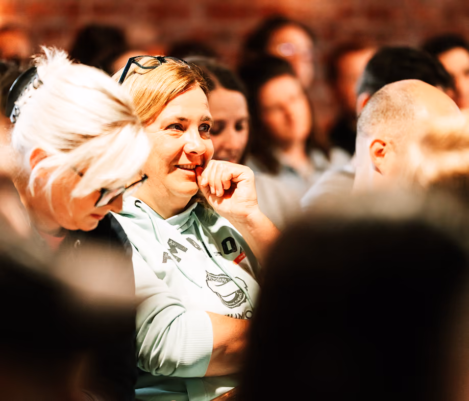 Frau mit blondem Haar und hellgrünem Hoodie lächelt in einem Publikum bei einer Veranstaltung.