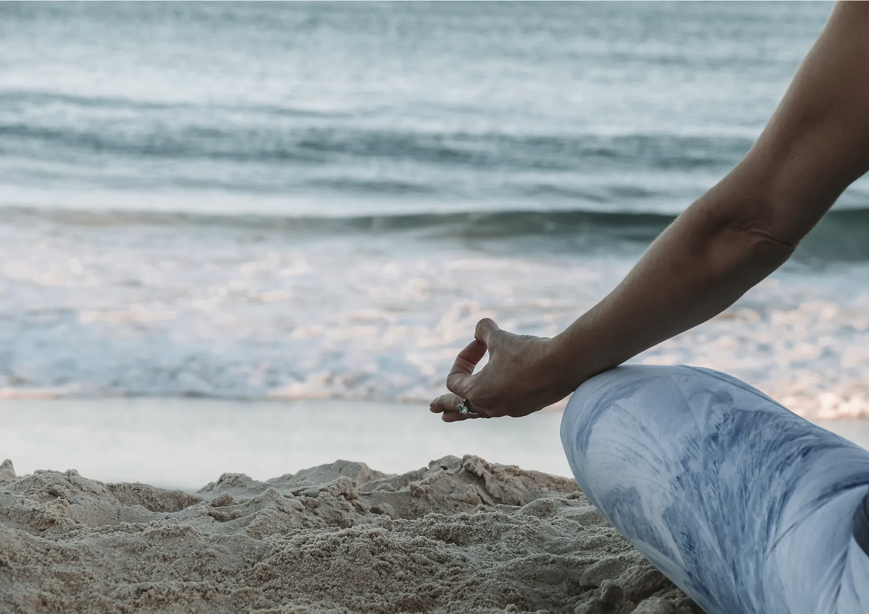 Person wearing blue leggings sitting cross-legged on sandy beach near ocean waves, meditating with hand gesture.