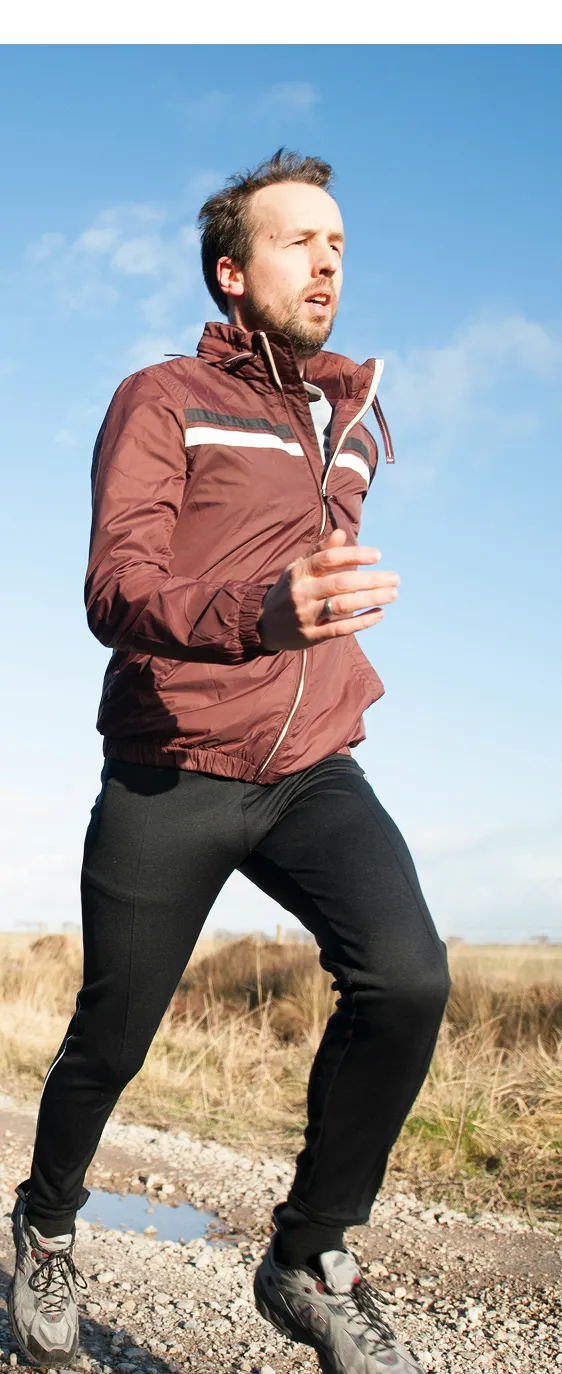 Man jogging outdoors on rocky path wearing maroon jacket, black pants, and gray running shoes under blue sky.