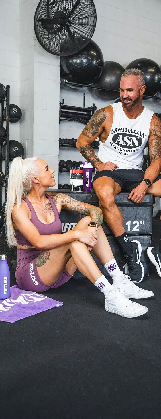 Two tattooed fitness enthusiasts in activewear sitting in a gym with ASN sports nutrition products.
