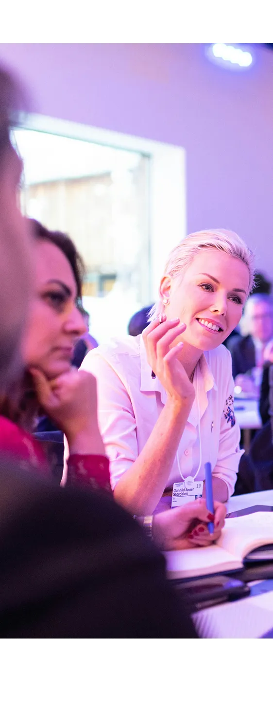 Woman in white shirt talking and gesturing while holding a pen at a group meeting.